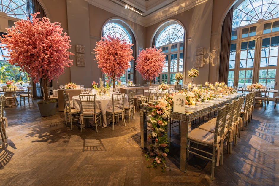 A spacious, elegant event hall illuminated by natural light streaming through large, arched windows, featuring three tall, lush pink cherry blossom trees in dark planters positioned along the walls. T