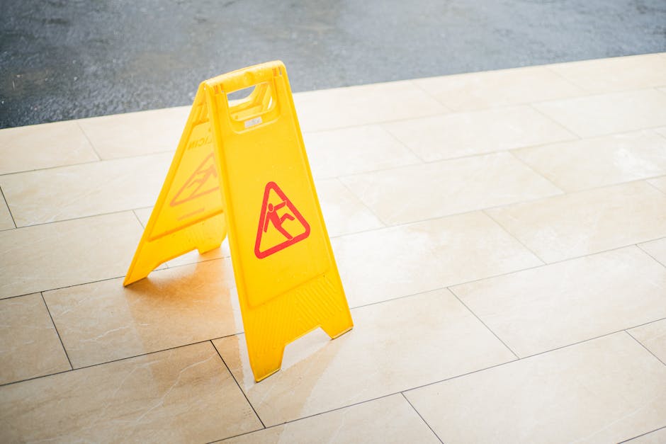 A yellow, freestanding wet floor warning sign with a red triangular symbol featuring a person slipping, placed on a beige tiled floor near the edge of the pavement outside a building. The tiles have a
