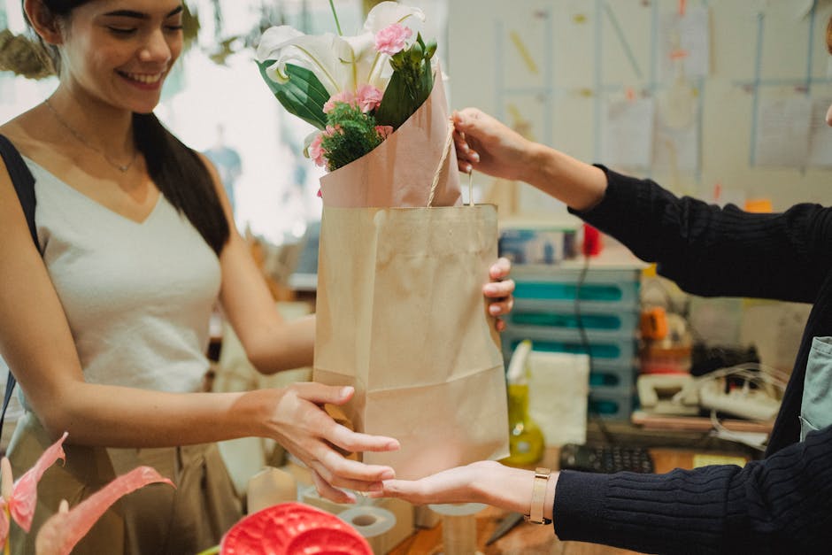 A woman with long dark hair, wearing a light-colored sleeveless top, is smiling as she receives a bouquet of fresh flowers from a florist in a workshop. The bouquet features white lilies, pink roses, 