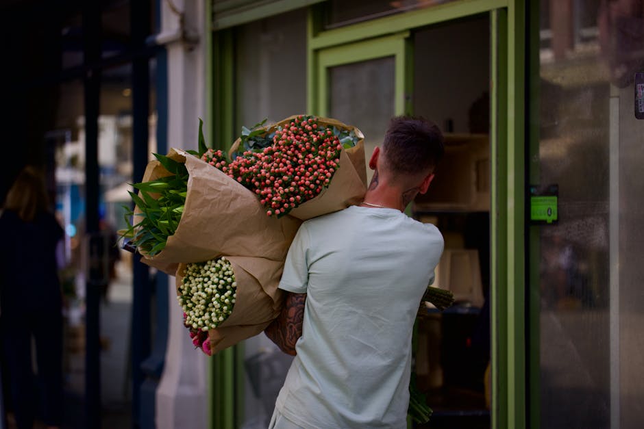 A person with short dark hair, wearing a light grey t-shirt, is holding a large bouquet of fresh flowers wrapped in brown paper. The bouquet contains pink and white flowers, possibly hypericum berries