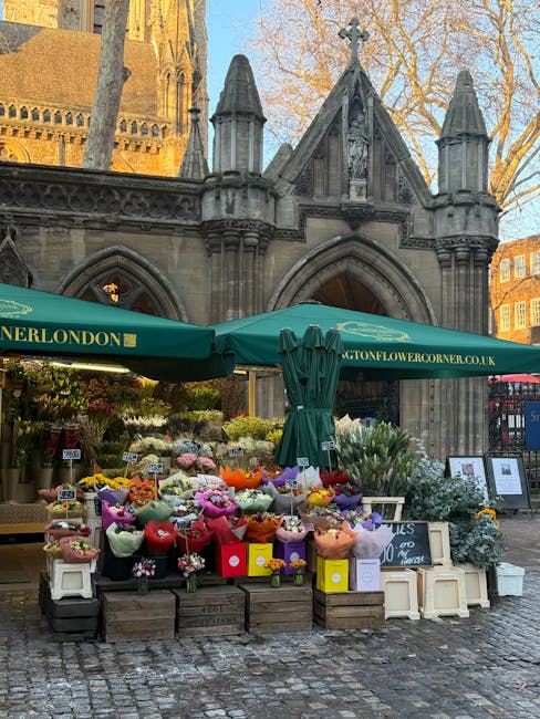 A vibrant flower stall situated outdoors in front of a historic stone building with Gothic architectural details at Kensington Gardens. The stall is covered by a large green canopy bearing the website