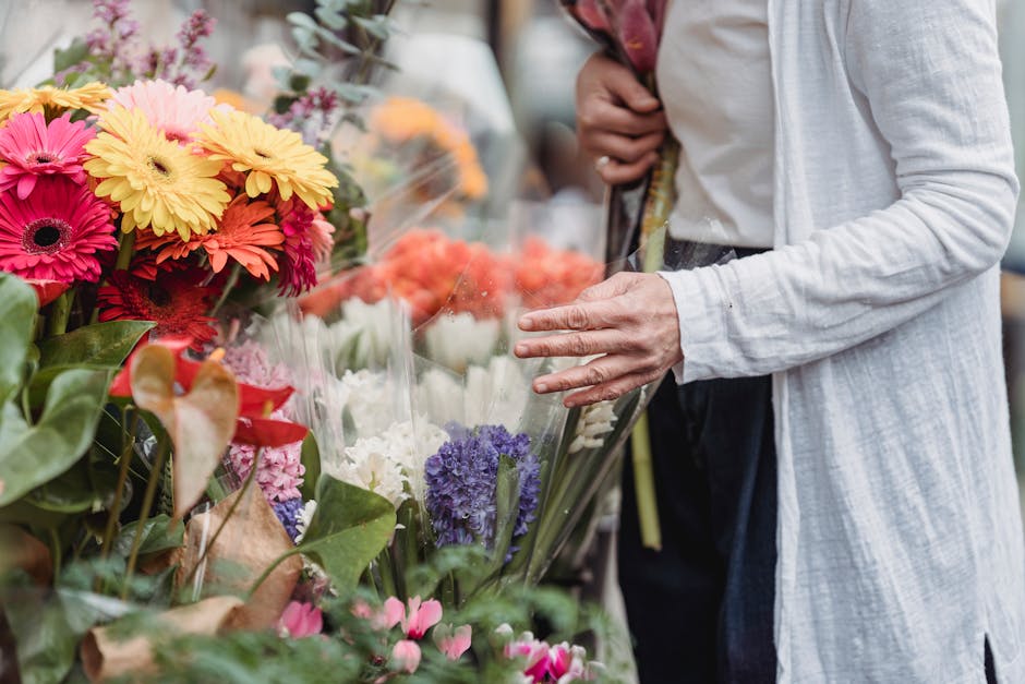 A person in a light grey cardigan and dark trousers is selecting a floral arrangement from a vibrant display of fresh flowers at a florist shop near Kensington Gardens. The bouquet includes bright pin