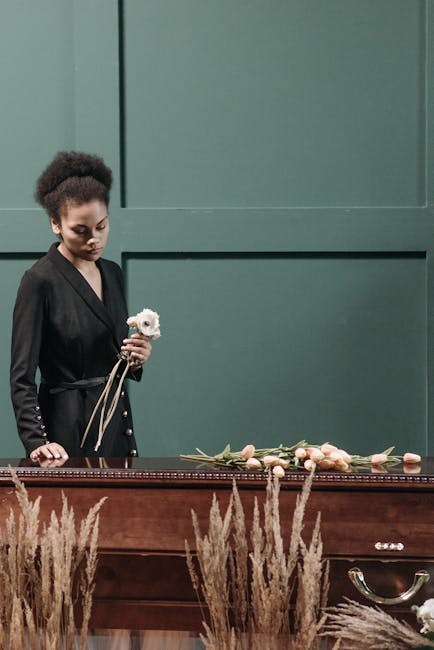 A woman with dark hair styled in a natural, textured updo, dressed in a sophisticated black blazer, stands beside a wooden casket adorned with a floral arrangement. She holds a white flower in her rig