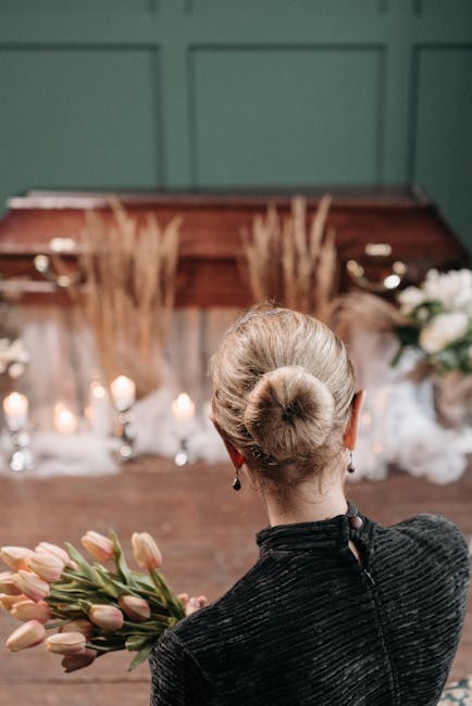 A woman with her blonde hair styled in a classic bun, dressed in a dark, textured top, is holding a bouquet of pink tulips with green stems and leaves. She stands indoors with a wooden backdrop decora