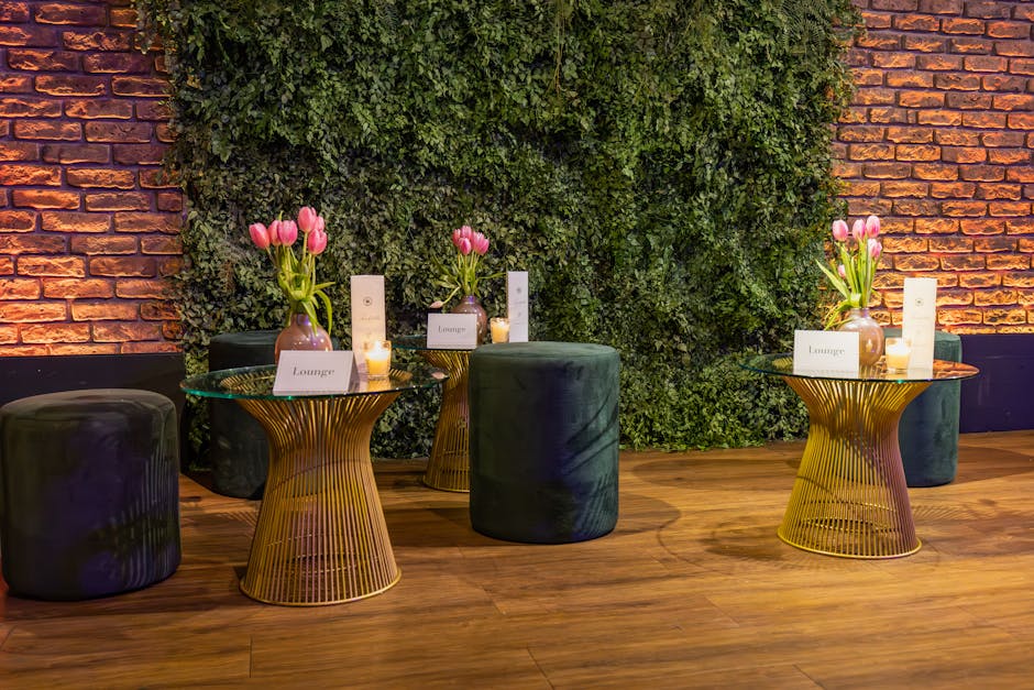 Three floral arrangements featuring pink tulips in small, rounded, light purple vases placed on metallic gold wireframe tables with glass tops. The tables are situated in a stylish lounge area with a