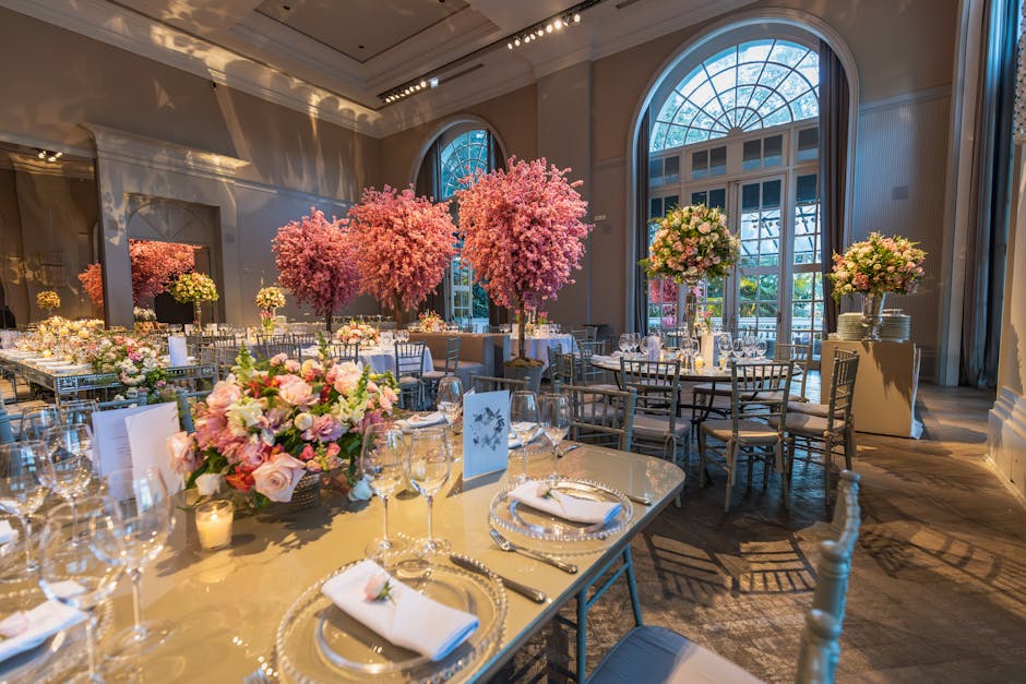 Elegant indoor wedding floral arrangement with large, vibrant pink and blush cherry blossom trees positioned near tall, arched windows, filling the room with natural light. The tables are decorated wi